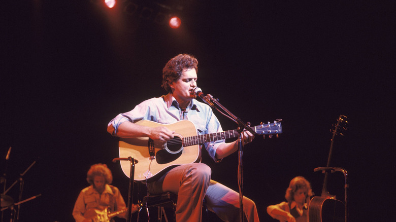 Harry Chapin playing guitar and singing while sitting on stage