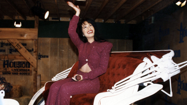 Selena Quintanilla Pérez smiles and waves to the crowd as she rides in a white carriage wearing a red outfit