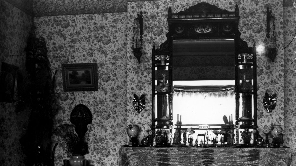 The mantlepiece of a Victorian suburban villa, crowded with ornaments and backed by poisonous wallpaper. 