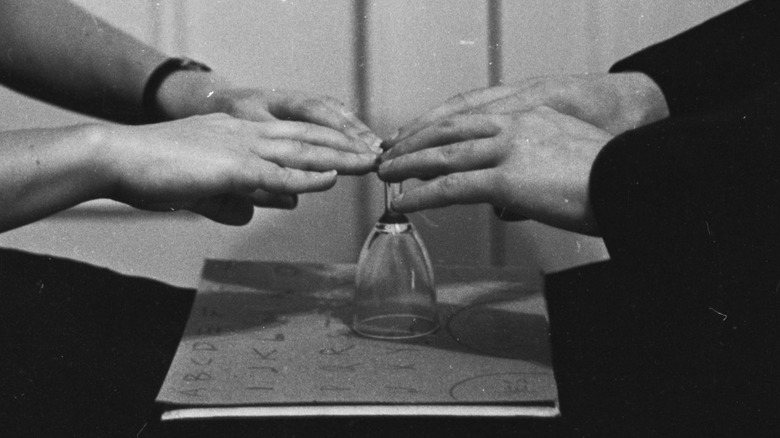 Two hands guide a modified Ouija board with a glass instead of a planchette