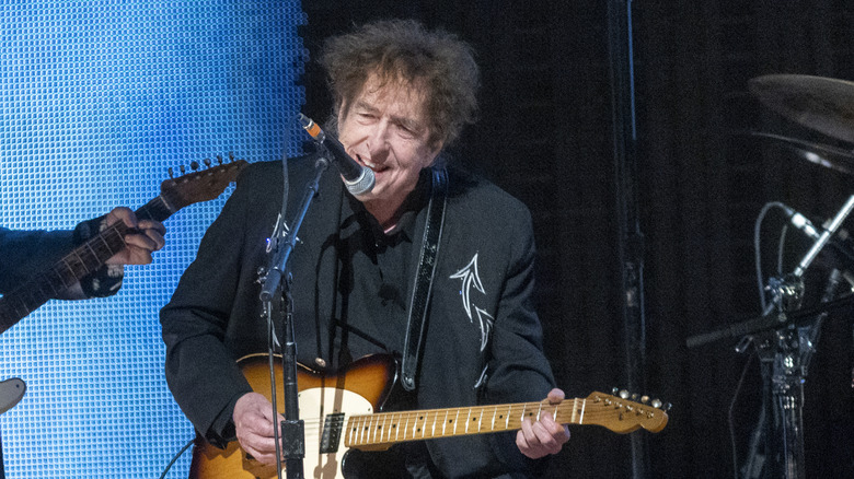 Bob Dylan on stage in dark outfit with microphone and guitar