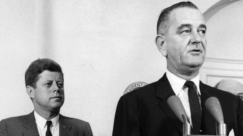 John F Kennedy stands behind Lyndon Johnson
