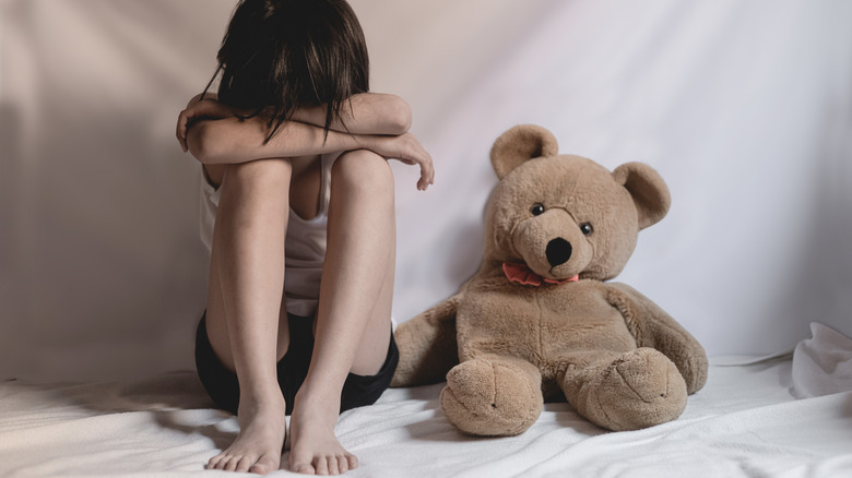 girl with head down sitting next to teddy bear