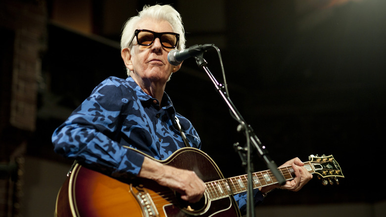 Nick Lowe wearing sunglasses playing guitar in fron t of a microphone