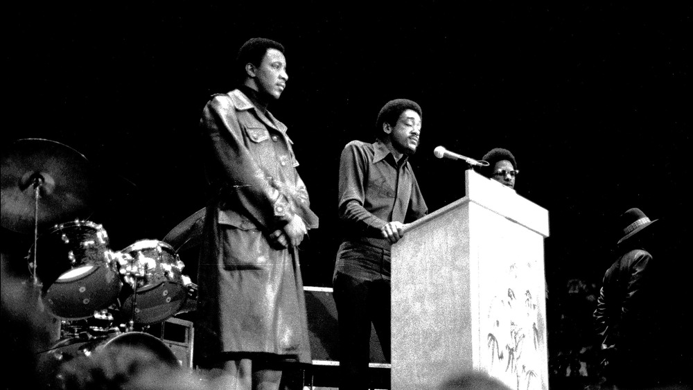 Black Panther Party Leader Bobby Seale speaking at podium on stage at the John Sinclair Freedom Rally at the Crisler Arena, surrounded by bodyguards, on December 12, 1971