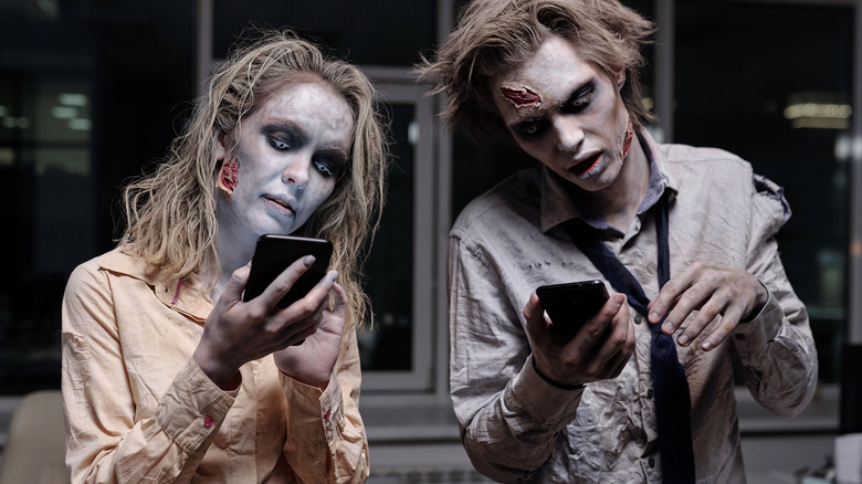 Dead and spooky businesspeople with zombie greasepaint on their faces and hands using smartphones