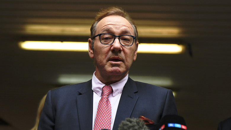 Kevin Spacey in suit and glasses speaking to reporters