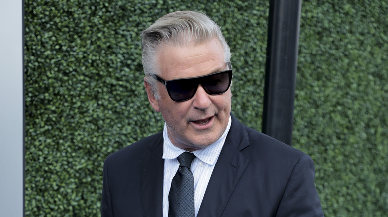 Alec Baldwin wearing sunglasses suit outside by hedge