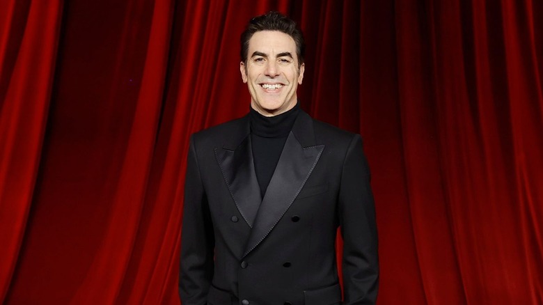 Sacha Baron Cohen stood smiling black suit red curtains behind him