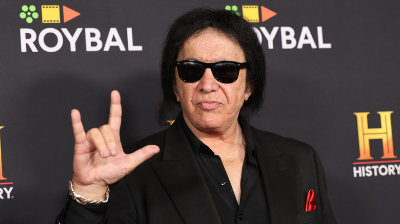 Gene Simmons in black outfit and sunglasses doing devil horns salute