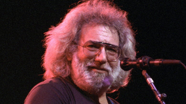 An older Jerry Garcia smiling at camera stands at microphone, smiling and playing guitar.