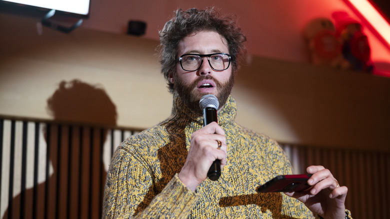 T.J. Miller yellow sweater glasses holding microphone