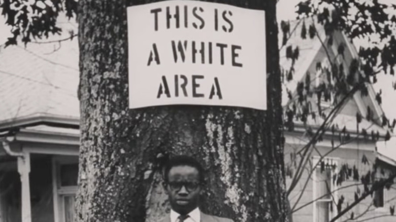 Black and white photo of a black man standing under a sign: "This is a white area"