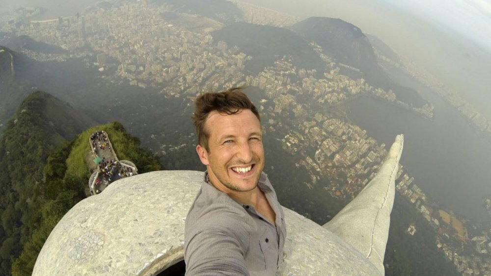 dangerous social media photo, view of a man hoisting a camera above his head while standing on a tall stone statue overlooking a city and forest