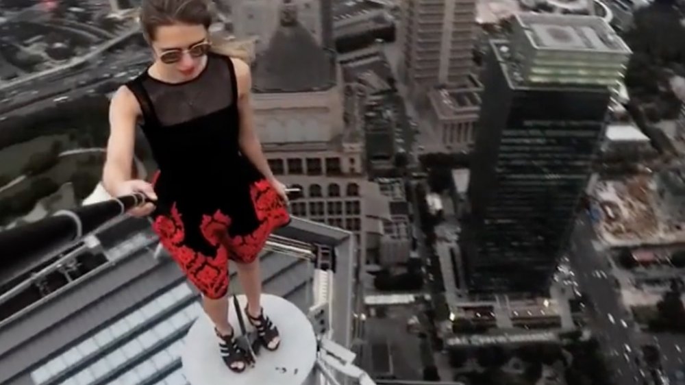 dangerous social media photo, young woman in black and red dress on small high ledge holding selfie stick