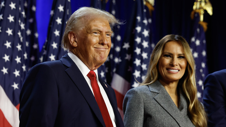 Donald and Melania Trump smiling onstage in front of flags