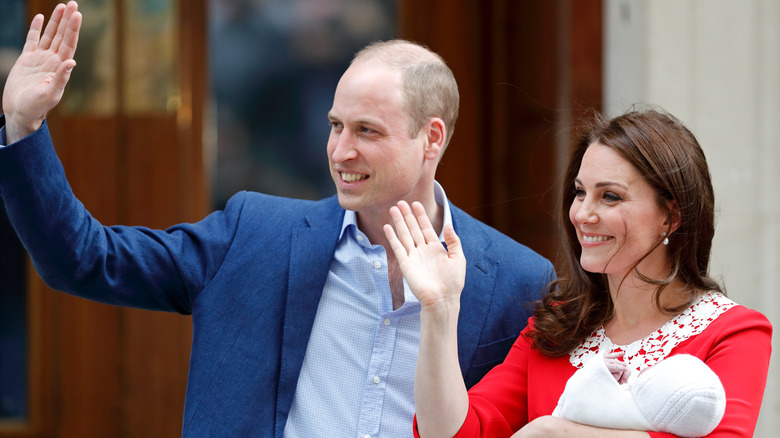 Prince William in suit and Kate Princess of Wales in red dress waving with Prince Louis