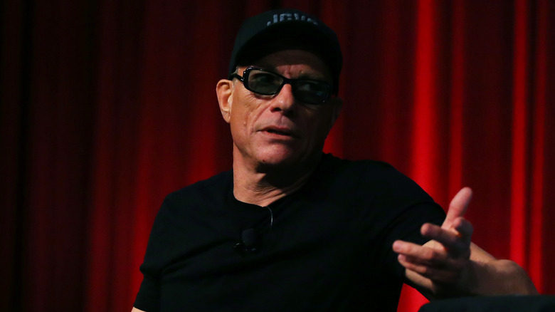 Jean-Claude Van Damme gesturing in black cap sunglasses