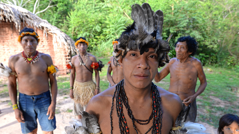 Awá tribesmen with feathered headresses in front of hut