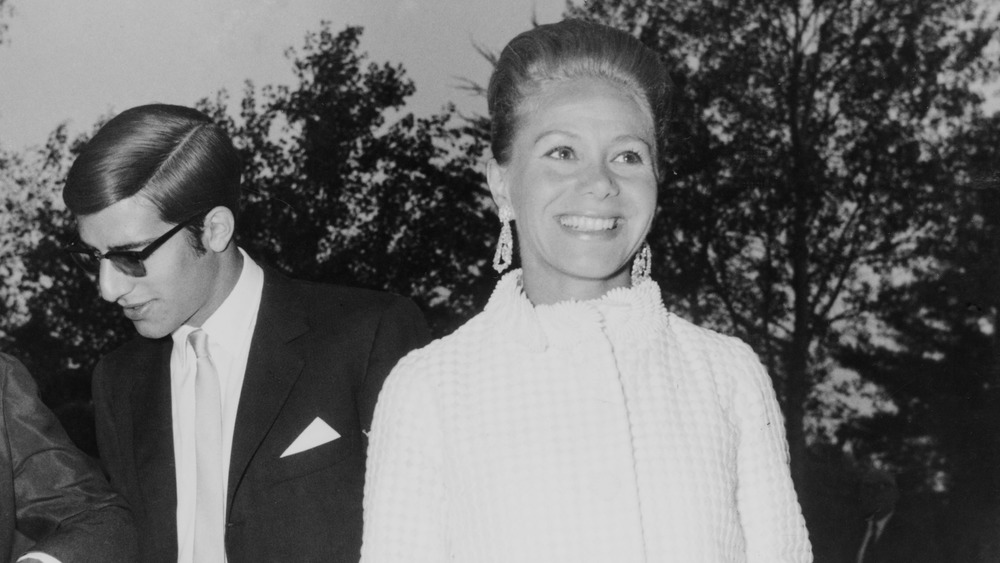 Tina Onassis Niarchos standing and smiling with her son Alexander Onassis behind her in conversation