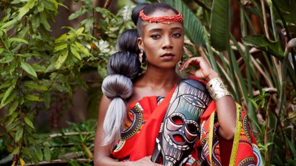 Portrait of a beautiful young African woman wearing traditional clothing and holding a mask while sitting outside with foliage in the background