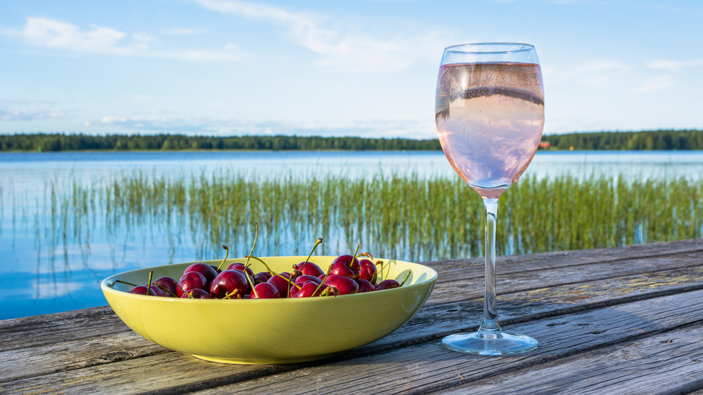 Glass of rose and a bowl of cherries on a wooden table overlooking a reed-strewn lake