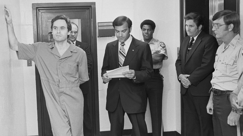 Ted Bundy leans against wall wearing  jail jumpsuit as sheriff reads indictment