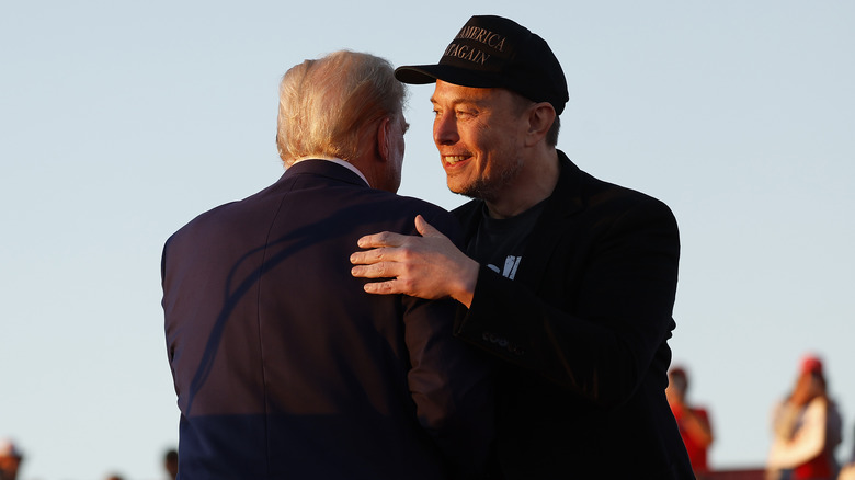 Elon Musk in dark baseball cap hugging Donald Trump on stage