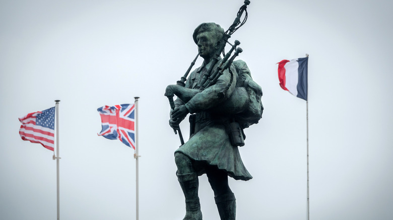 Bill Millin Memorial on Sword Beach in Normandy, with flags and statue of Bill Millin playing bagpipes.