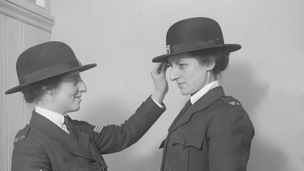 A woman police sergeant helps her companion get the tilt of her uniform hat right. Thy have both been appointed for duty at a munitions works (