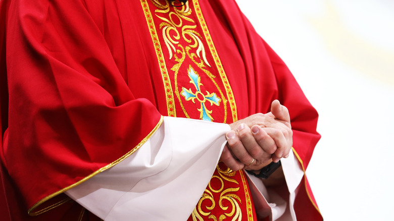 Person in red priest robes gold design hands clasped