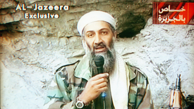 Osama Bin Laden speaks into microphone next to gun