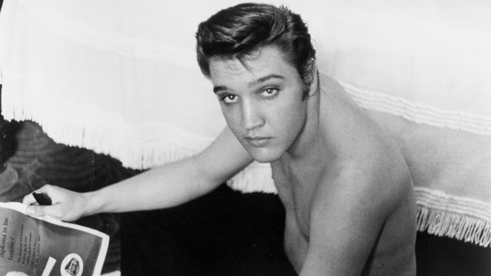 Elvis Presley poses for a portrait while reading a magazine sitting next to a bed in 1956