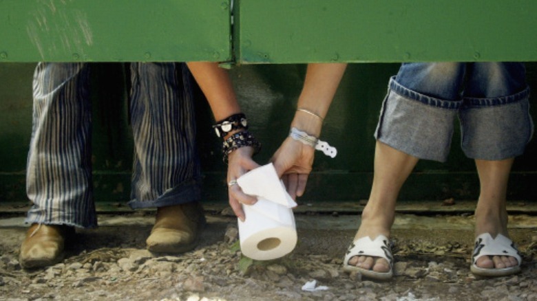 Two people in bathroom stalls pass a roll of toilet paper 