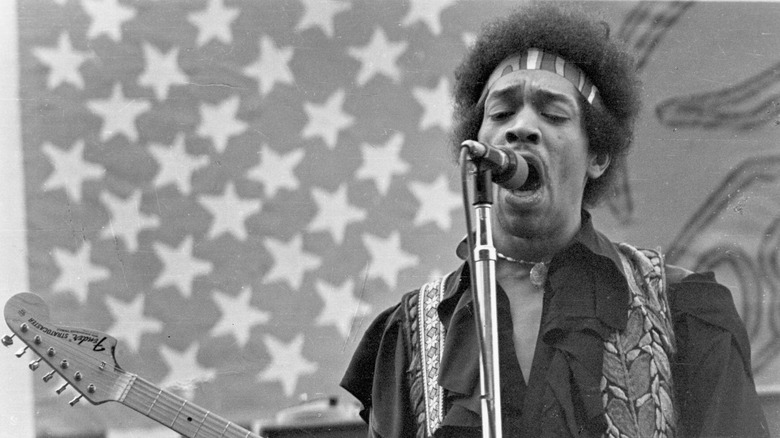 Jimi Hendrix singing and playing guitar with an American flag backdrop