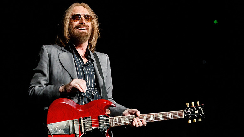 Tom Petty smiles on stage while wearing sunglasses and a grey suit and playing a red guitar