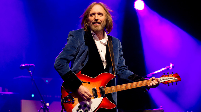 Tom Petty performs on stage with a guitar in his hands and purple lights in the background