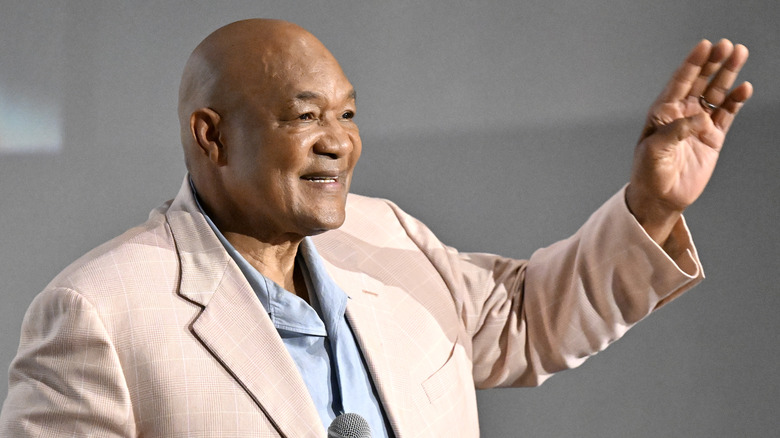 George Foreman in a blue shirt and cream colored jacket waving